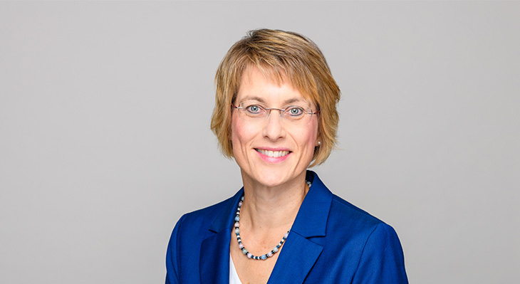 Frau mit kurzen blonden Haaren, Brille und blauer Jacke l&auml;chelt vor grauem Hintergrund