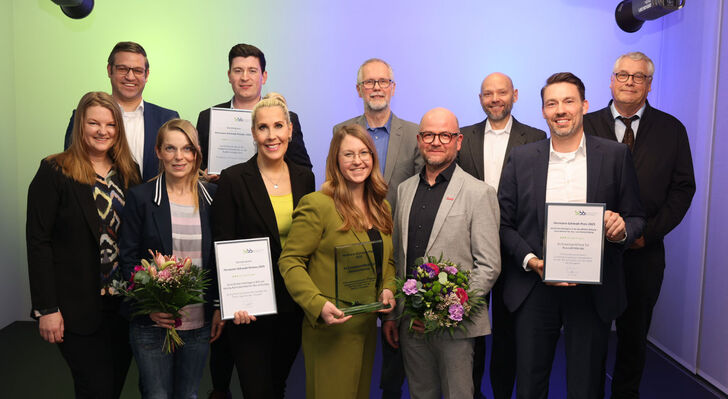 Eine Gruppe von elf Personen in formeller Kleidung auf einer B&uuml;hne, einige halten Urkunden und Blumenstr&auml;u&szlig;e