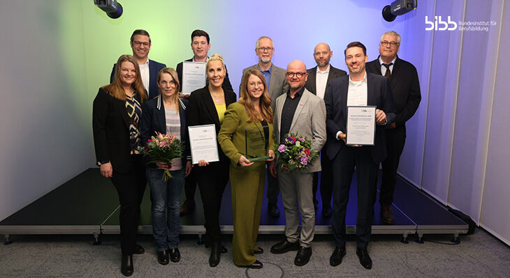 Eine Gruppe von elf Personen in formeller Kleidung auf einer B&uuml;hne, einige halten Urkunden und Blumenstr&auml;u&szlig;e