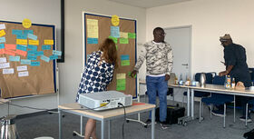 Three people are standing in a meeting room in front of two noticeboards covered in colourful notes; a table with a projector and drinks is in the foreground