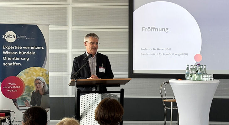 Herr Ertl steht auf einer B&uuml;hne vor einem Rednerpult und begr&uuml;&szlig;t die Teilnehmenden der Veranstultung.