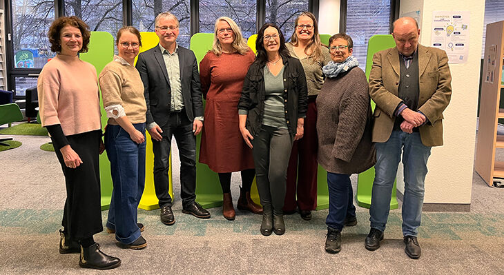  Eine Gruppe von acht Personen steht in einem modernen B&uuml;ro mit gro&szlig;en Fenstern und gr&uuml;nen Sitzm&ouml;beln im Hintergrund