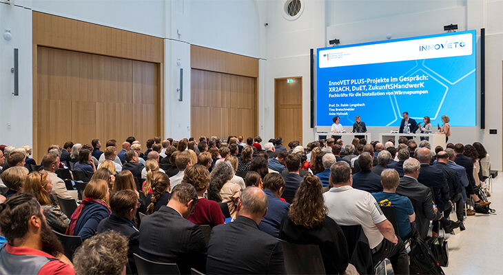 Publikum sitzt in einem Konferenzraum und blickt auf eine B&uuml;hne