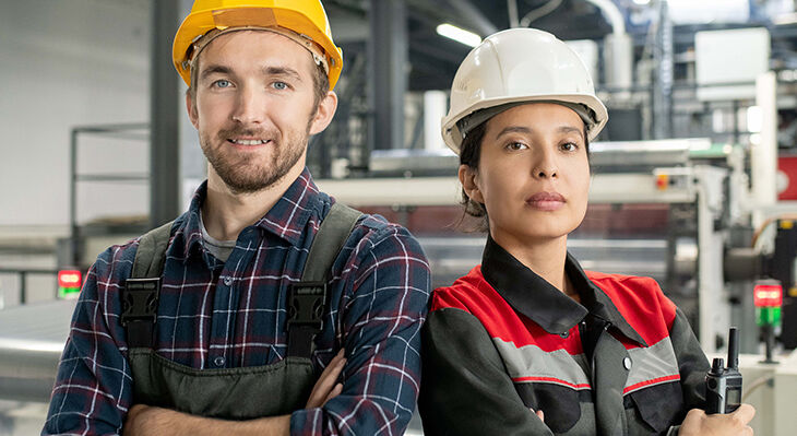 Zwei Bauerbeiter/-innen verschr&auml;nken vor einer Fabrikhalle die Arme und gucken in die Kamera. 