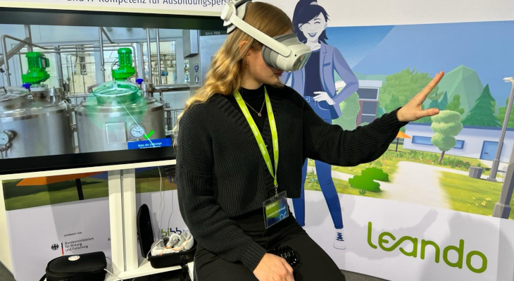 Frau auf einer Messe mit Virtual Reality Headset. Im Hintergrund sieht man auf einem Bildschirm eine Fabrik.
