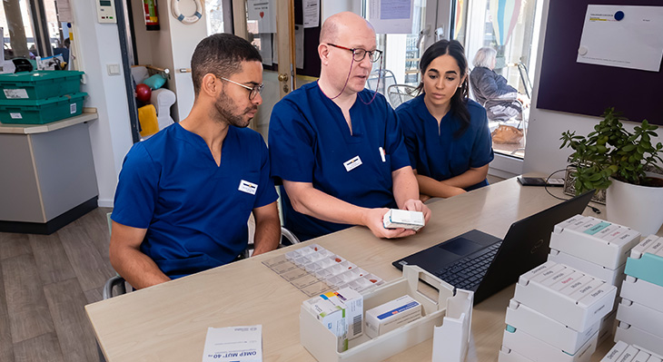 Es ist ein Pfleger zu sehen mit zwei Auszubildenden, die bei der Tablettenvergabe eingewiesen werden. Die drei Menschen sitzen vor einem Tisch mit vielen Tablettendosen