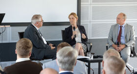 Drei Personen in Anzug und Kleid sitzen auf St&uuml;hlen bei einer Podiumsdiskussion, eine Person h&auml;lt ein Mikrofon und gestikuliert