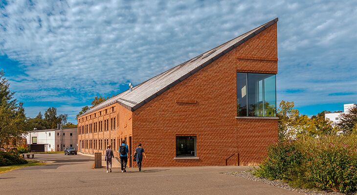 Ungleichm&auml;&szlig;ig gebautes Steinhaus mit gro&szlig;er Fensterfront, davor gehen 3 Personen.