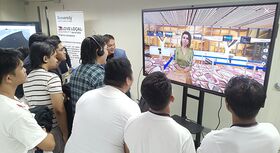 A group of people stand in front of a large screen showing a virtual person.