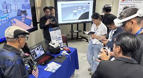 Several people at an exhibition stand with monitors displaying 3D models and technical illustrations.