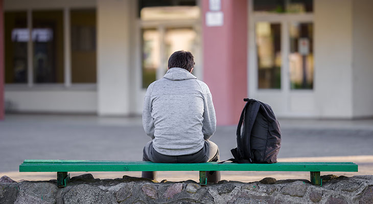 Jugendlicher sitzt mit dem Rücken zum Betrachter auf einer grünen Bank, neben sich einen Rucksack stehend