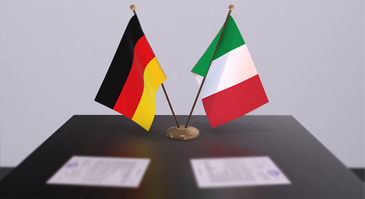 Tischflaggen von Deutschland und Italien auf einem Schreibtisch, mit verschwommenen Urkunden im Vordergrund, die symbolisch für die Unterzeichnung der vereinbarten Zusammenarbeit stehen