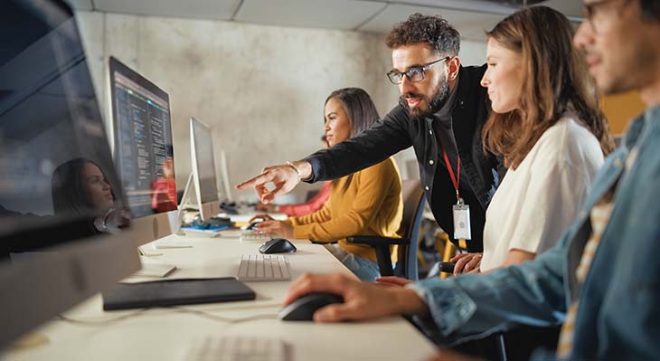 Drei Personen sitzen vor laptops. Hinter den drei Personen steht eine Person die etwas auf einem der Laptops zeigt.