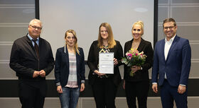 F&uuml;nf Personen stehen nebeneinander vor einer grauen Wand, eine Person h&auml;lt eine Urkunde, eine andere einen Blumenstrau&szlig;.