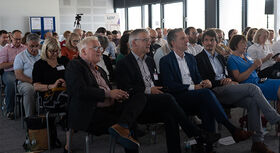 Gruppe von Menschen sitzt in einem hellen Raum bei einer Veranstaltung und h&ouml;rt aufmerksam zu.