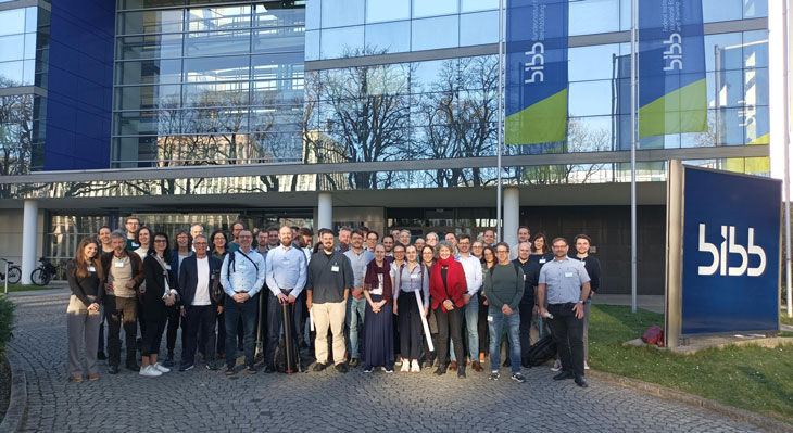 Gruppe von Menschen steht vor einem modernen Glasgeb&auml;ude mit zwei Fahnen und einem gro&szlig;en Schild mit der Aufschrift 'bibb'