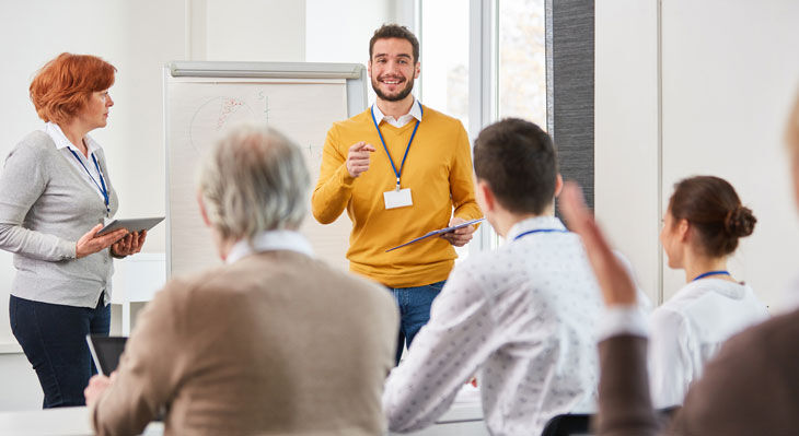 Mann in gelbem Pullover pr&auml;sentiert vor Gruppe sitzender und stehender Personen in hellem Seminarraum