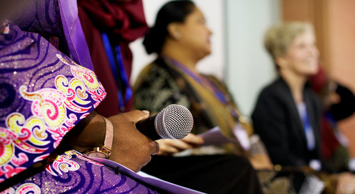 Nahaufnahme einer Person in farbenfroher Kleidung, die ein Mikrofon h&auml;lt, im Hintergrund unscharfe Personen in Sitzreihen