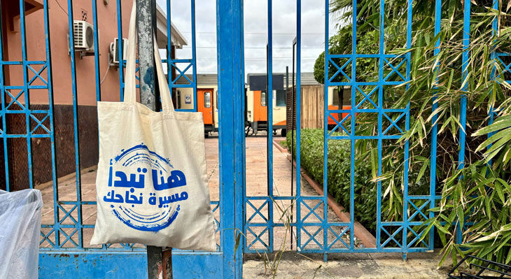 Stoffbeutel mit arabischem Schriftzug h&auml;ngt an einem blauen Metalltor vor einem Bahnsteig mit abgestellten Waggons