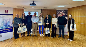 Gruppe von neun Personen steht in einem Raum mit Holzw&auml;nden vor zwei aufgestellten Bannern und h&auml;lt wei&szlig;e Stofftaschen mit blauem Aufdruck