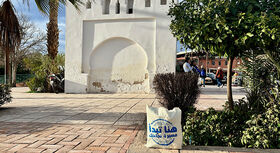 Beutel mit arabischer Schrift steht auf gepflastertem Platz vor wei&szlig;er Mauer mit Bogen und umgeben von B&uuml;schen