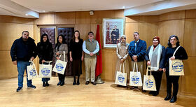 Gruppe von neun Personen steht in einem Raum mit Holzboden vor einer Wand mit einem gro&szlig;en Portr&auml;t, jede Person h&auml;lt eine Stofftasche mit arabischer Schrift