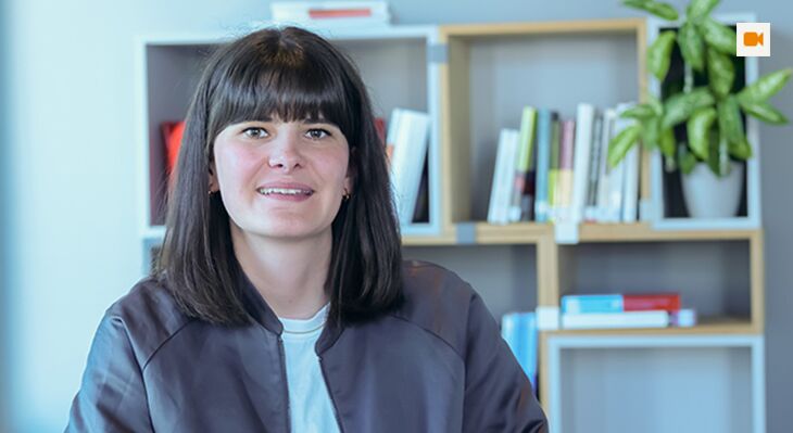 Frau mit schulterlangen dunklen Haaren sitzt l&auml;chelnd vor einem B&uuml;cherregal in einem hellen Raum