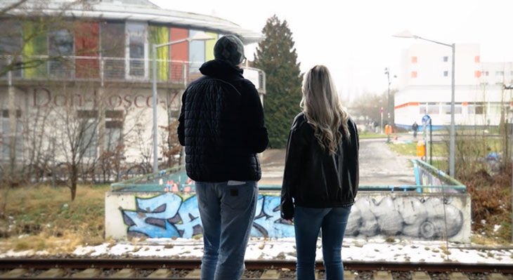 Zwei Personen stehen mit dem R&uuml;cken zur Kamera vor einem Graffiti auf einer niedrigen Mauer und blicken auf ein Geb&auml;ude mit bunten Fenstern.