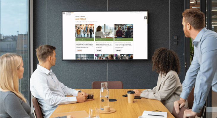 Vier Personen sitzen an einem Konferenztisch und schauen auf einen Bildschirm an der Wand, auf dem eine Webseite mit mehreren Kacheln angezeigt wird.