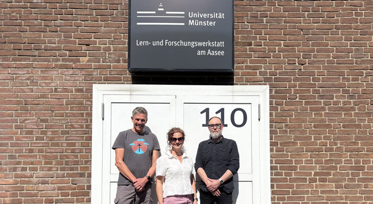 Drei Personen stehen vor einer wei&szlig;en T&uuml;r mit der Nummer 110 an einer Backsteinwand, dar&uuml;ber ein Schild mit dem Schriftzug 'Universit&auml;t M&uuml;nster Lern- und Forschungswerkstatt am Aasee'