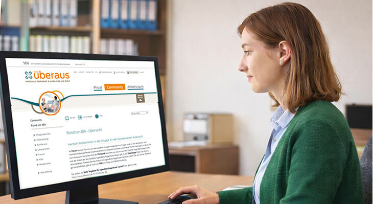 Eine Frau  mit gr&uuml;nem Pullover sitzt an einem Schreibtisch und blickt auf einen Monitor mit der Webseite "&uuml;beraus"