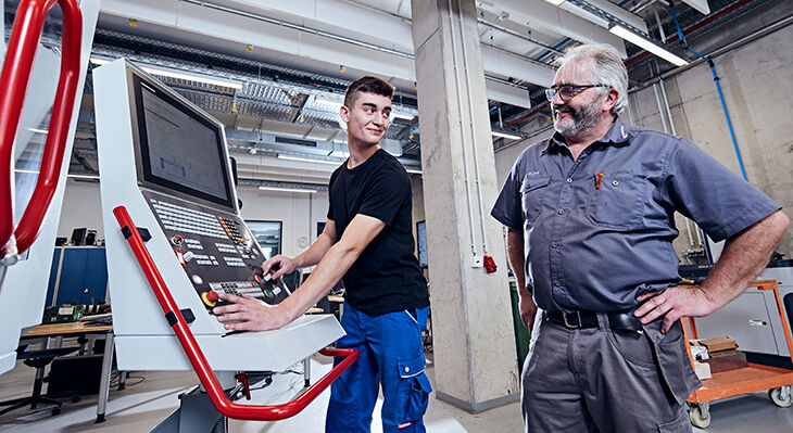 Junger Mann arbeitet an einer digitalen Maschine, neben ihm sein Ausbilder. Beide lächeln sich an.