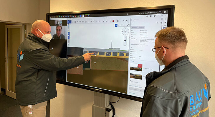 Der Mann links im Bild erklärt seinem Kollegen rechts im Bild an einem großen Monitor ein Gebäudemodell.