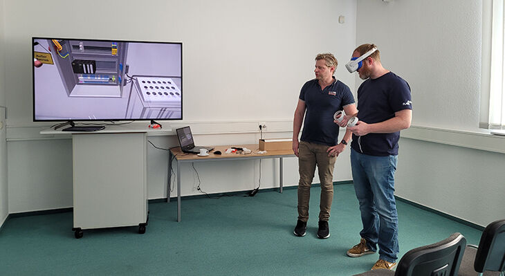 Links ein großer Monitor, rechts daneben stehen zwei Männer. Der vordere im Bild trägt eine VR-Brille.
