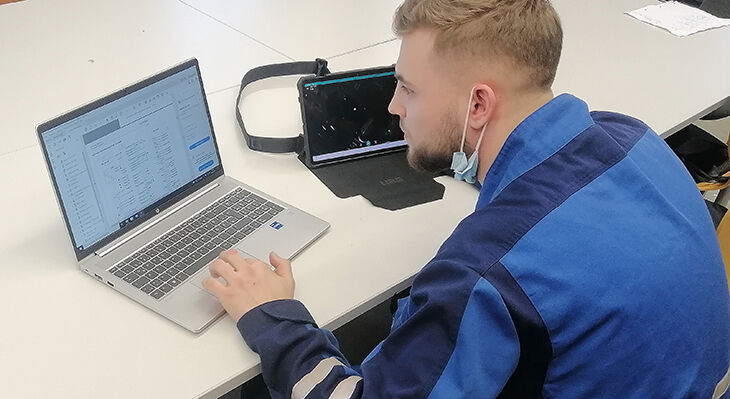 Ein junger, blonden Mann sitzt vor einem Laptop. Rechts neben dem Laptop steht ein weiterer, kleiner Bildschirm, auf dem ein Video live übertragen wird.