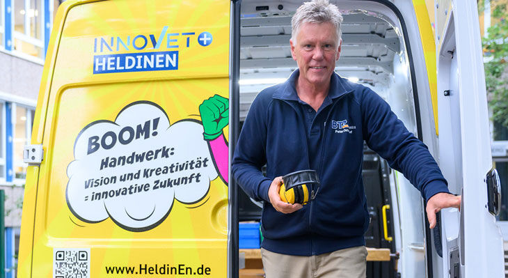 Mann mit blauem Poloshirt h&auml;lt Geh&ouml;rschutz vor einem wei&szlig;en Transporter mit gelber Werbefl&auml;che und Texten zu Handwerk und Innovation