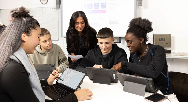 Gruppe von Jugendlichen sitzt an einem Tisch mit Tablets und Laptop, eine Frau steht dahinter und h&auml;lt Papiere