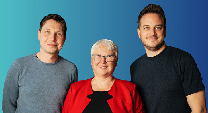 Drei Personen stehen vor einem zweifarbigen Hintergrund in Blau- und T&uuml;rkist&ouml;nen, die mittlere Person tr&auml;gt ein rotes Jackett, die anderen beiden tragen dunkle Oberteile