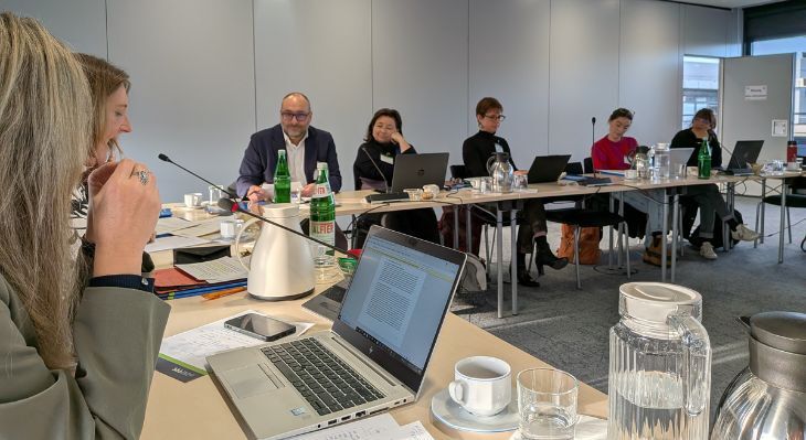 Mehrere Personen sitzen an einem langen Konferenztisch mit Laptops, Getr&auml;nken und Mikrofonen in einem  Besprechungsraum.