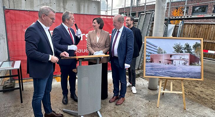 Pr&auml;sident N&uuml;nemann h&auml;lt eine Rede bevor die Zeitkapsel verbaut wird. Neben ihm stehen weitere Personen, darunter Staatssekret&auml;rin Wulf. Au&szlig;erdem zu sehen: das Schild zum geplanten Bau