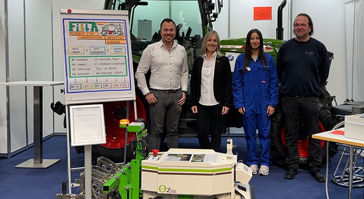 Das Team des Projektes FillA steht auf dem Messestand hinter dem Feldroboter. Dahinter befindet sich ein Traktor. 