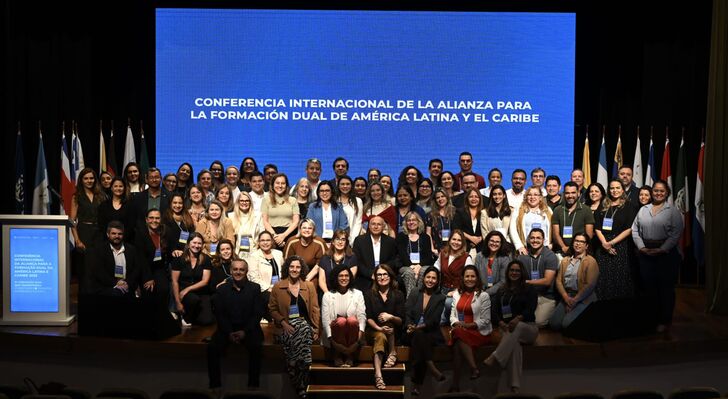 Group photo of all participants at the Alliance for Dual Education in Latin America and the Caribbean conference. Around 90 smiling people on stage, illuminated by spotlights.