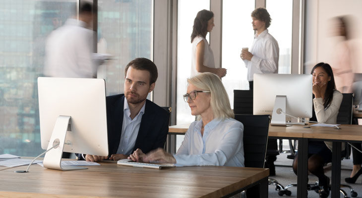 Zwei Personen sitzen an einem Schreibtisch und schauen gemeinsam auf einen Computerbildschirm, im Hintergrund stehen zwei weitere Personen im Gespr&auml;ch und eine Person sitzt an einem weiteren Schreibtisch mit Computer