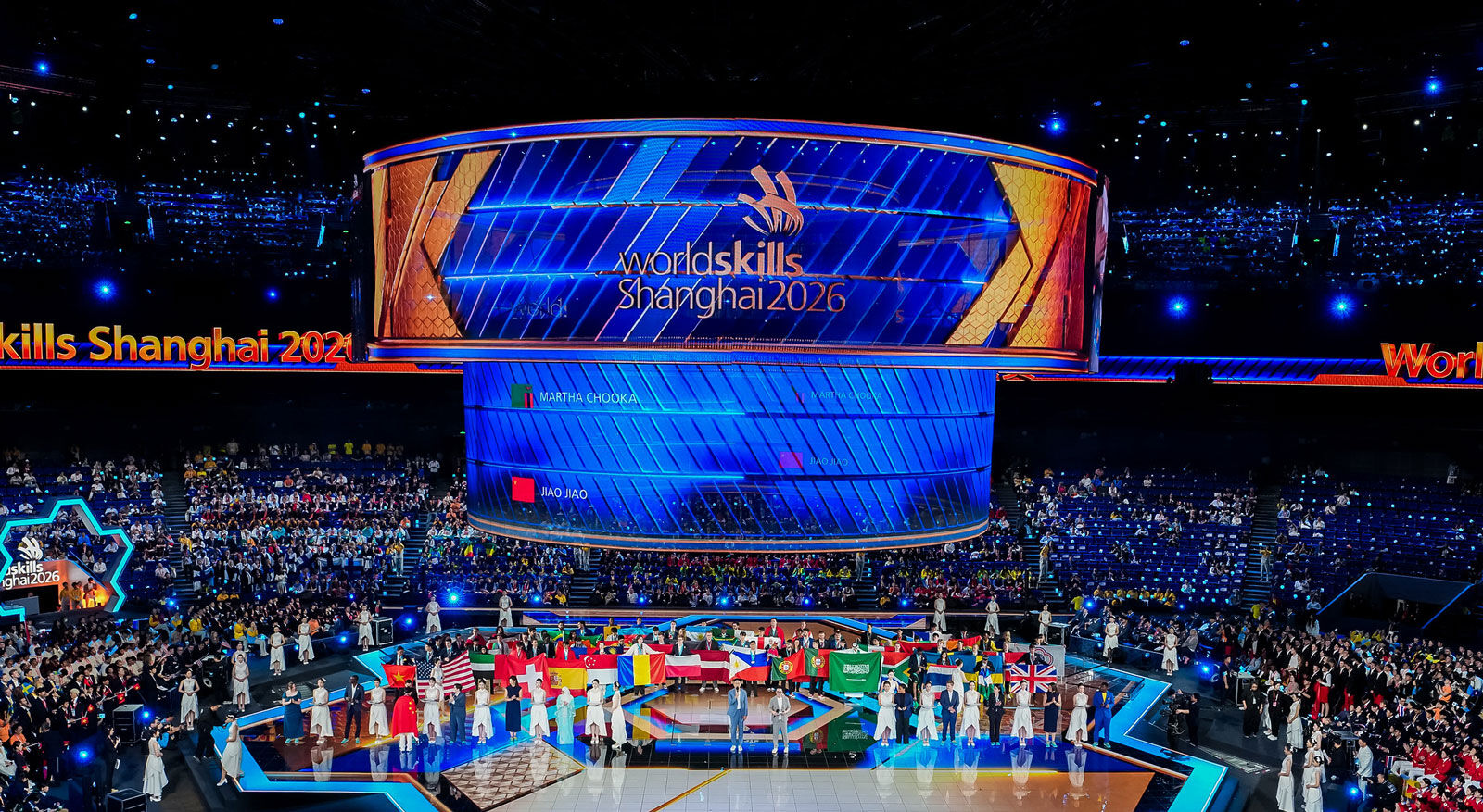 Menschen mit Fahnen stehen auf einer B&uuml;hne vor einer gro&szlig;en LED-Anzeige mit dem Schriftzug 'WorldSkills Shanghai 2022' in einer vollbesetzten Arena.