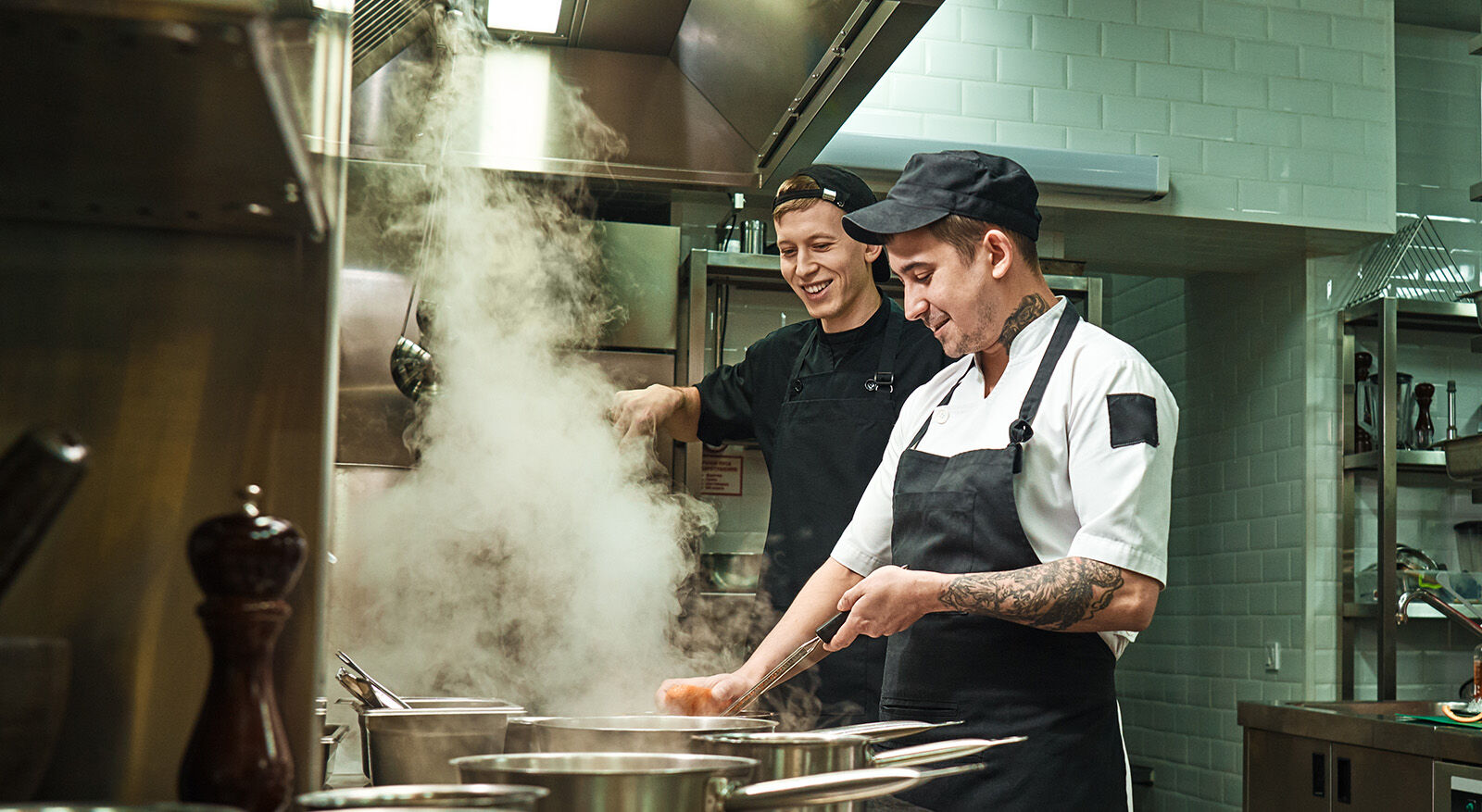 Zwei K&ouml;che in Kochkleidung arbeiten in einer professionellen K&uuml;che mit dampfenden T&ouml;pfen auf dem Herd
