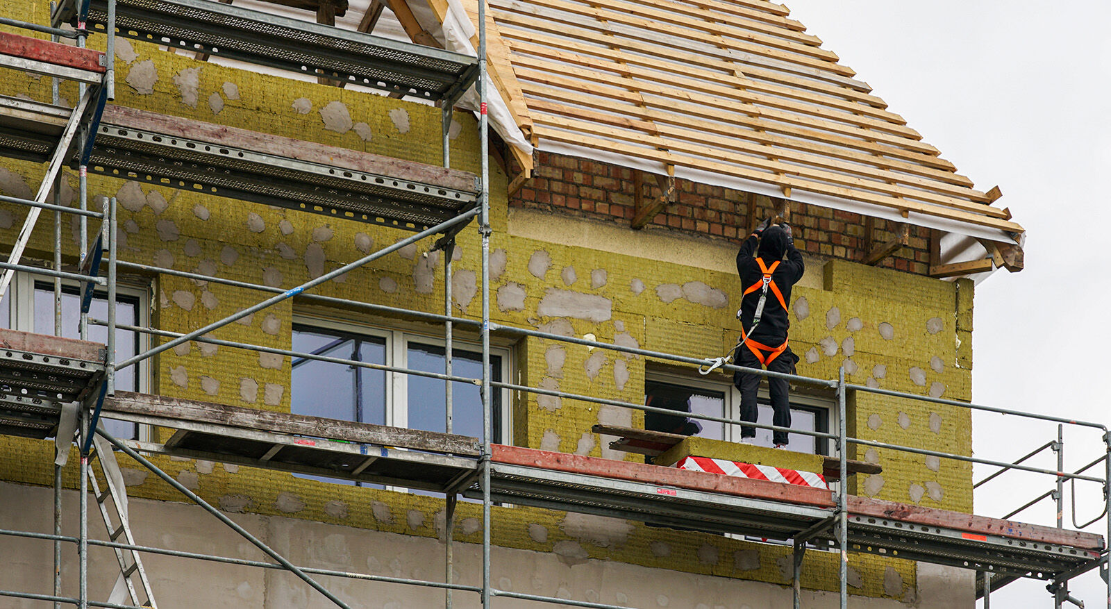 Arbeiter mit Sicherheitsgeschirr auf einem Ger&uuml;st vor einem teilweise mit D&auml;mmmaterial verkleideten Haus mit sichtbarem Dachstuhl
