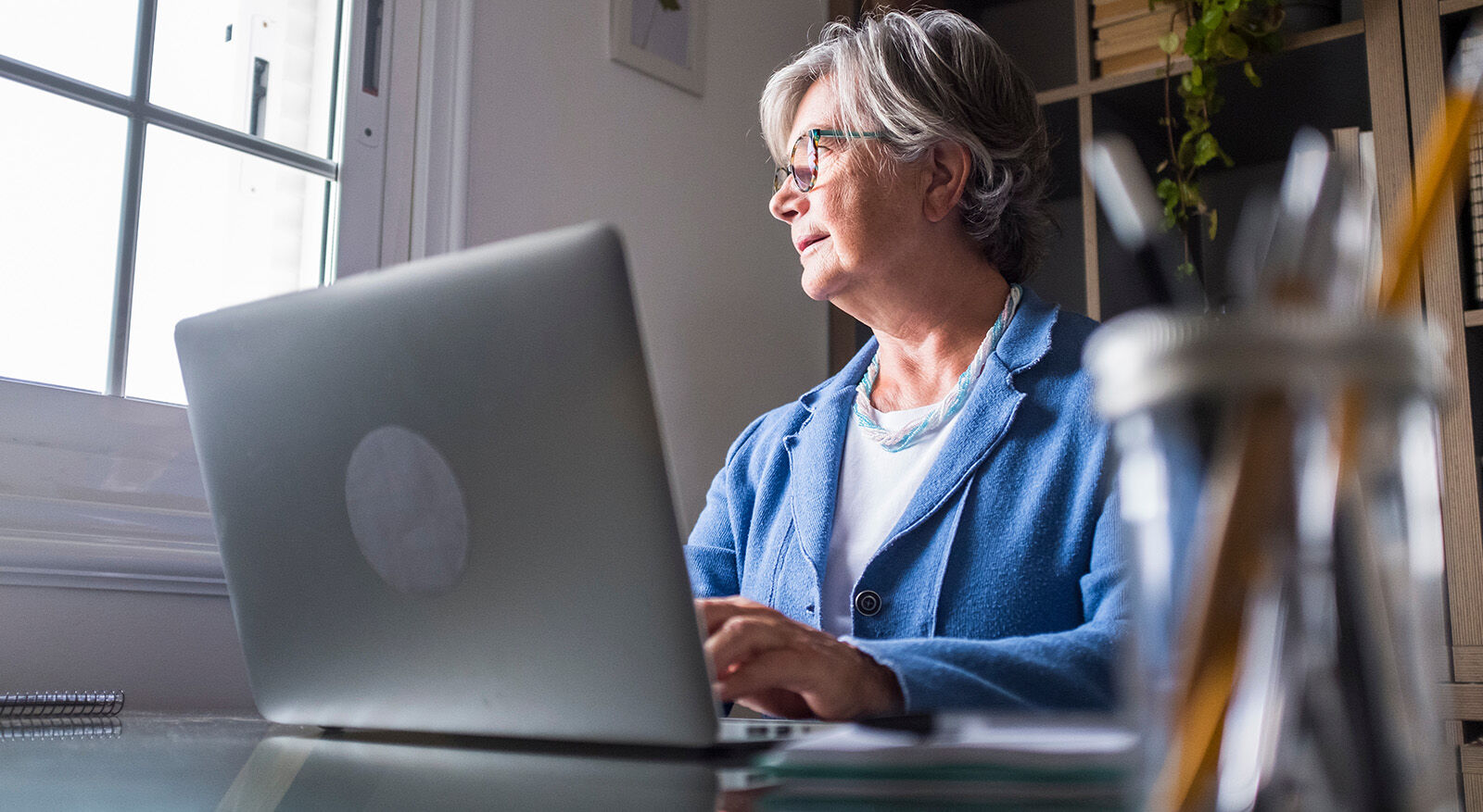 Eine ältere Frau schaut von Ihrem Laptop auf und sieht aus dem Fenster.