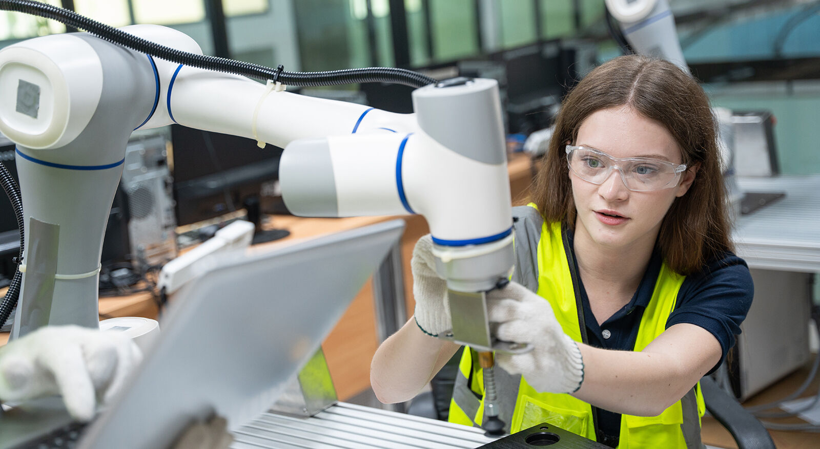 Frau mit Warnweste und Handschuhen bedient einen Industrieroboterarm in einer Fabrikumgebung