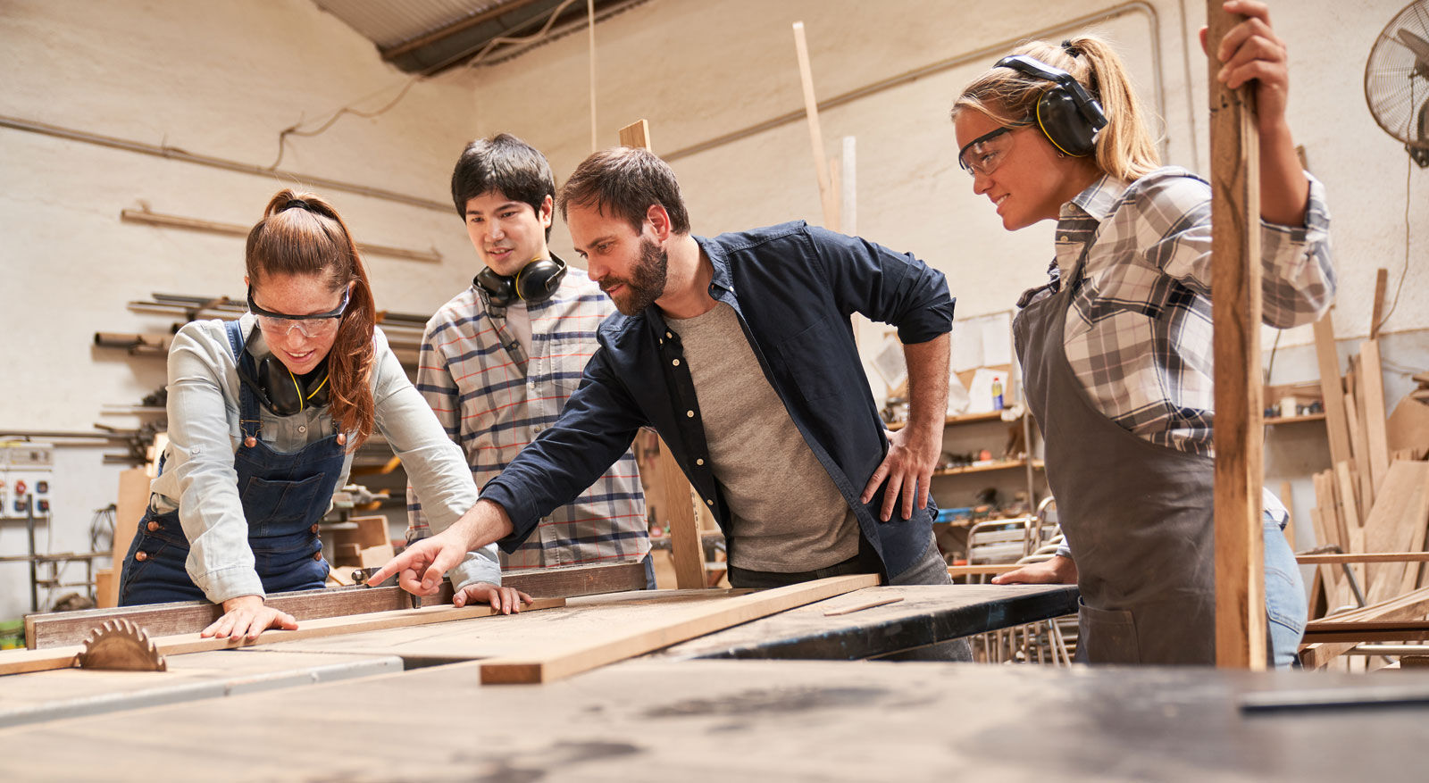 Vier Personen in Arbeitskleidung stehen an einem Holztisch in einer Werkstatt, eine zeigt auf ein Brett.