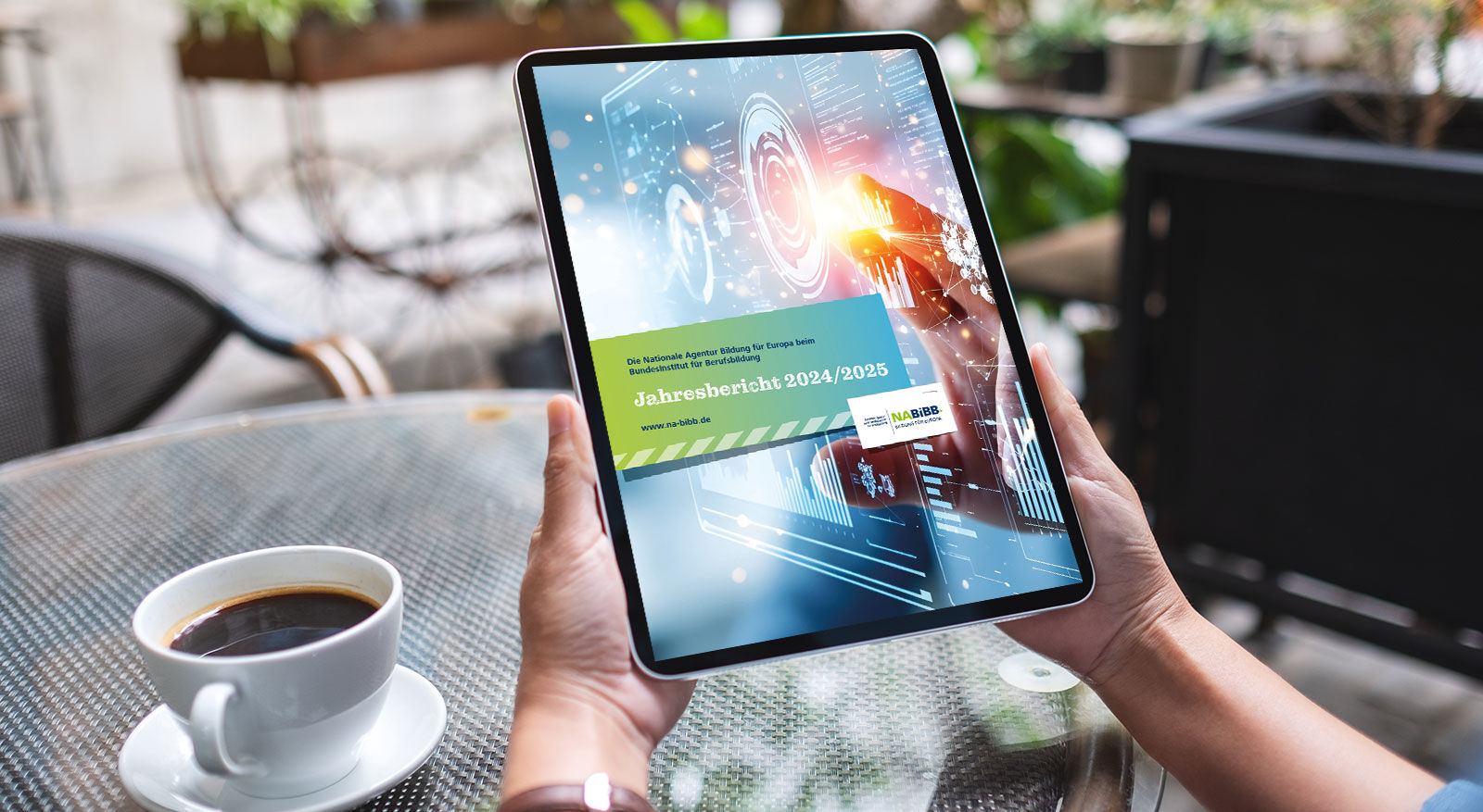 Person h&auml;lt Tablet mit digitaler Anzeige und Text 'Jahresbericht 2024/2025' vor einem Tisch mit Kaffeetasse